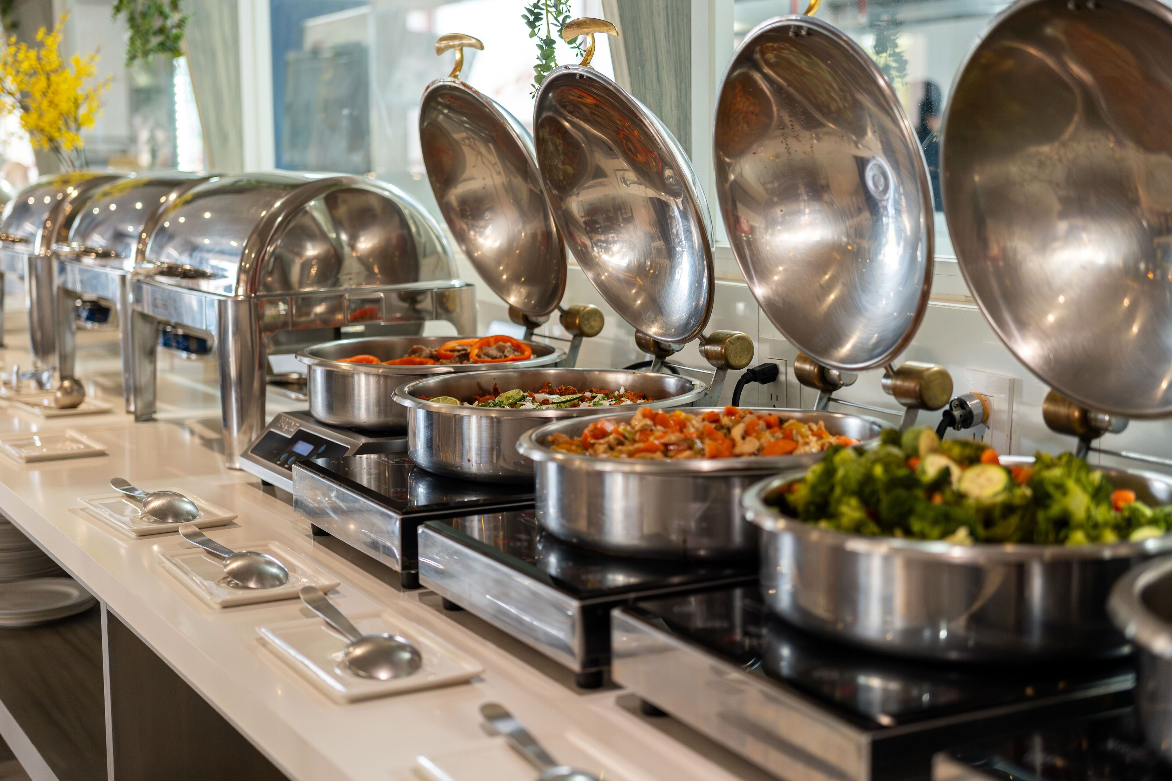 A buffet setup with multiple stainless steel serving dishes and utensils on a white countertop.