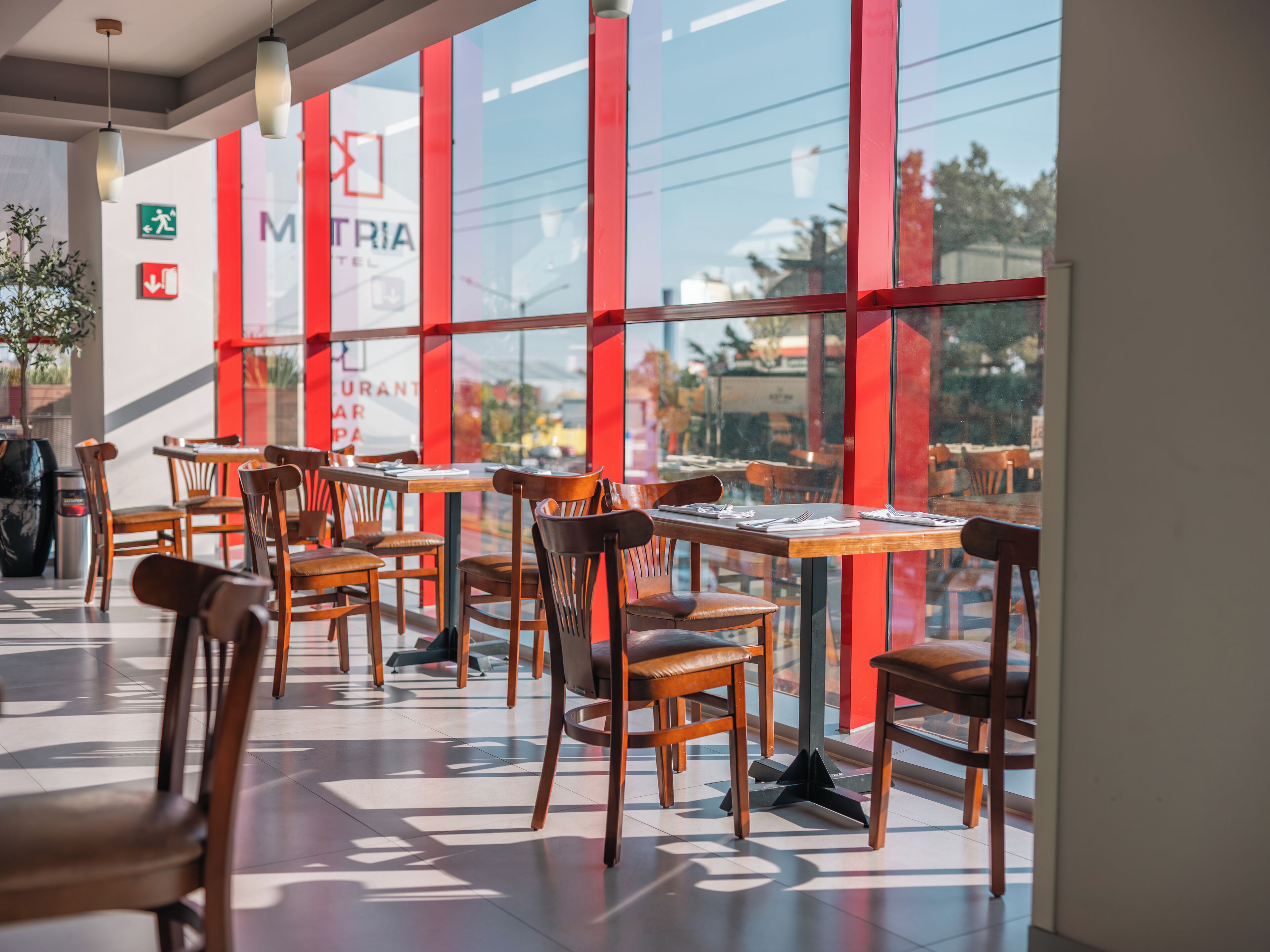 Un comedor interior con mesas y sillas de madera, ventanas amplias y elementos estructurales rojos.