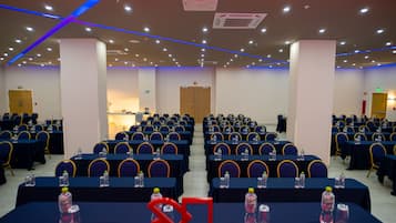 Un salón de conferencias con filas de mesas y sillas, equipado con botellas de agua y un proyector.