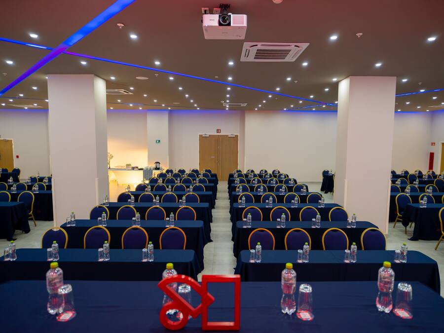 Un salón de conferencias con filas de mesas y sillas, equipado con botellas de agua y un proyector.