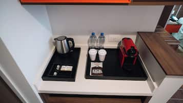Una kitchenette con cafetera, dos botellas de agua, dos tazas, un hervidor eléctrico rojo y un sobre de leche condensada.