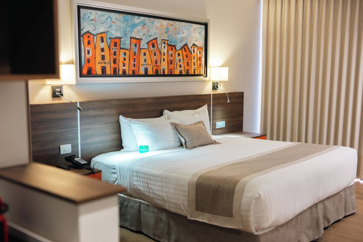 Una habitación de hotel con una cama grande, dos mesitas de noche, un buró, un cuadro sobre la cama y una ventana con cortinas.