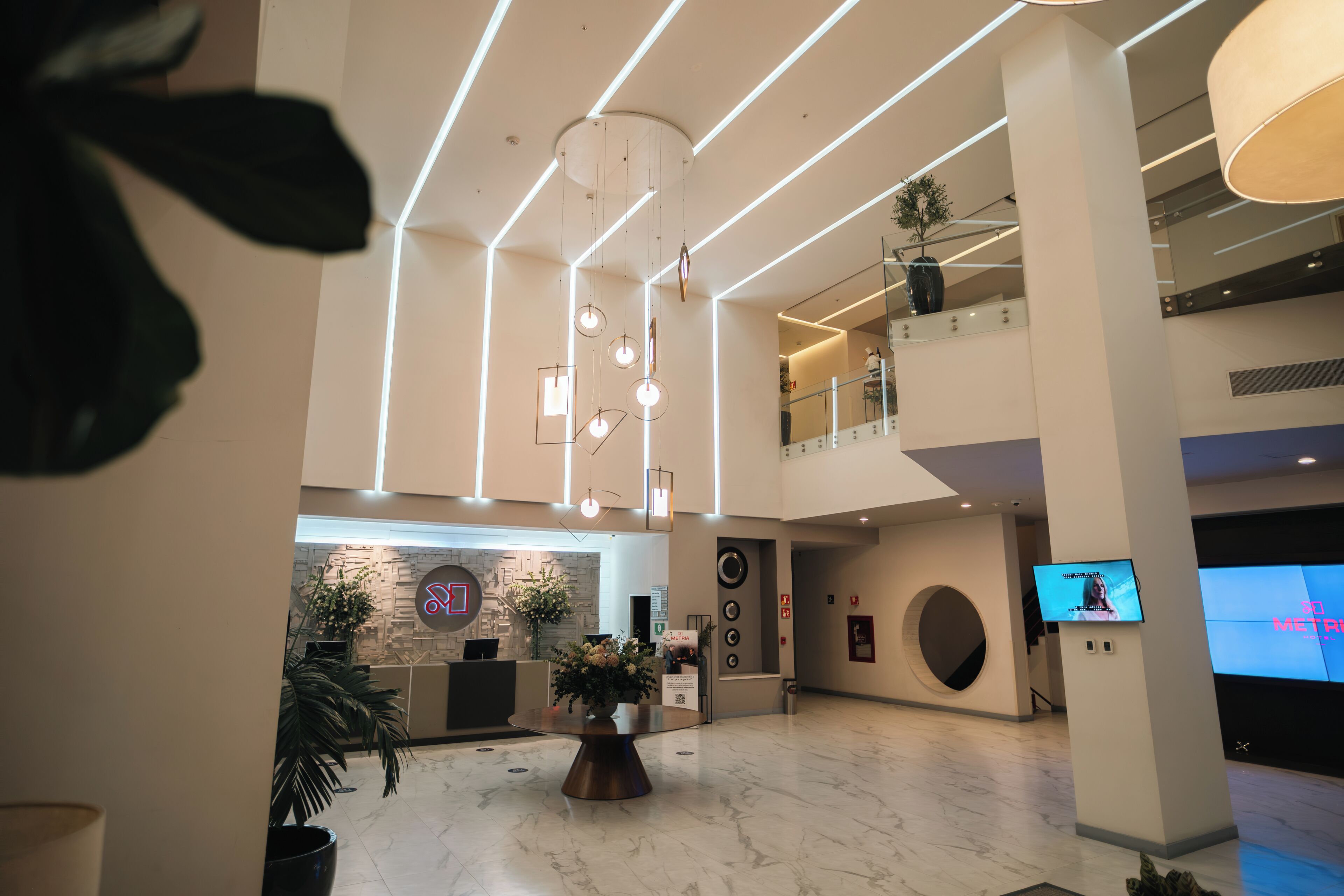 Un hall d’entrée spacieux avec un comptoir d’accueil, des plantes en pot et un grand écran diffusant des informations.