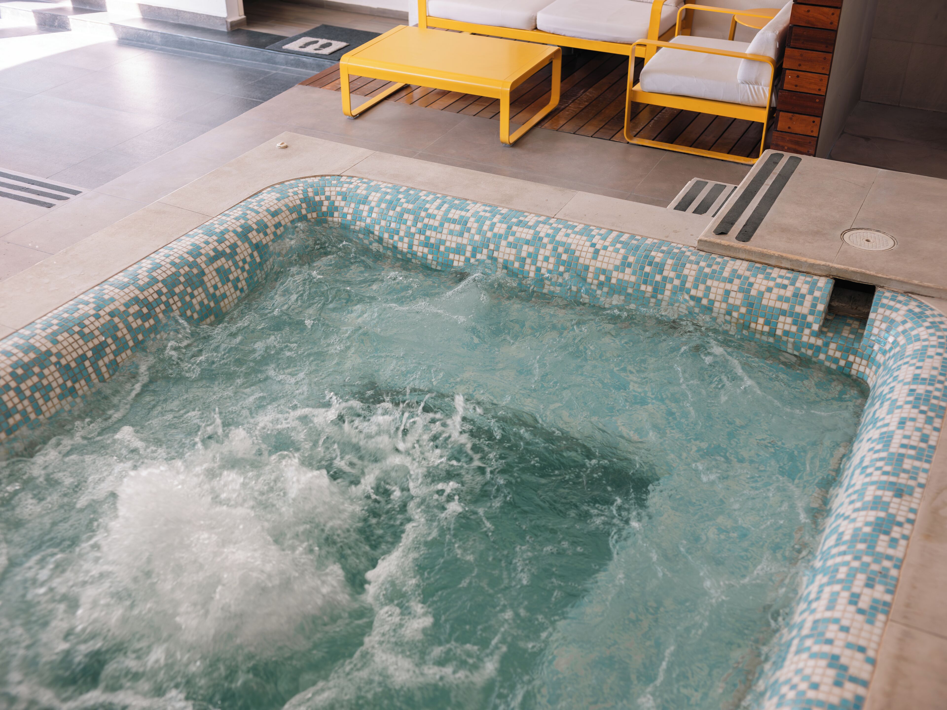 A hot tub with a tiled edge, surrounded by a wooden deck and yellow furniture.