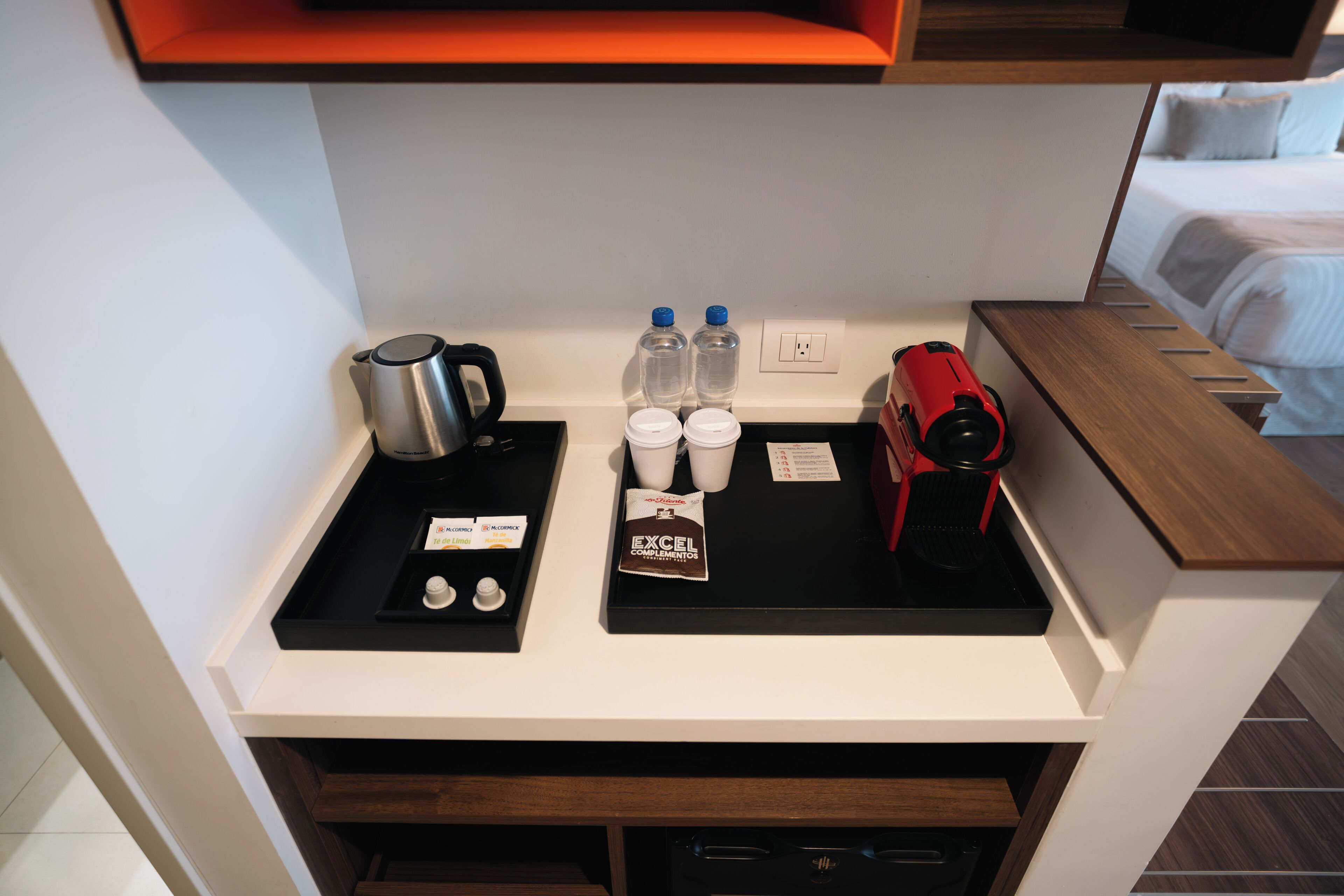 A hotel room's minibar with a coffee maker, two bottles of water, a tray with a coffee pod and a coffee cup, and a red electric kettle.