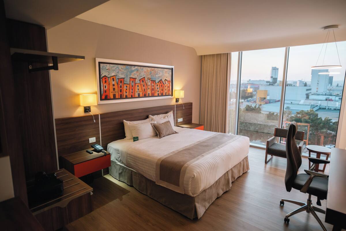 Habitación de hotel con una cama grande, un escritorio, una silla, una mesita de noche y vistas a la ciudad a través de la ventana.
