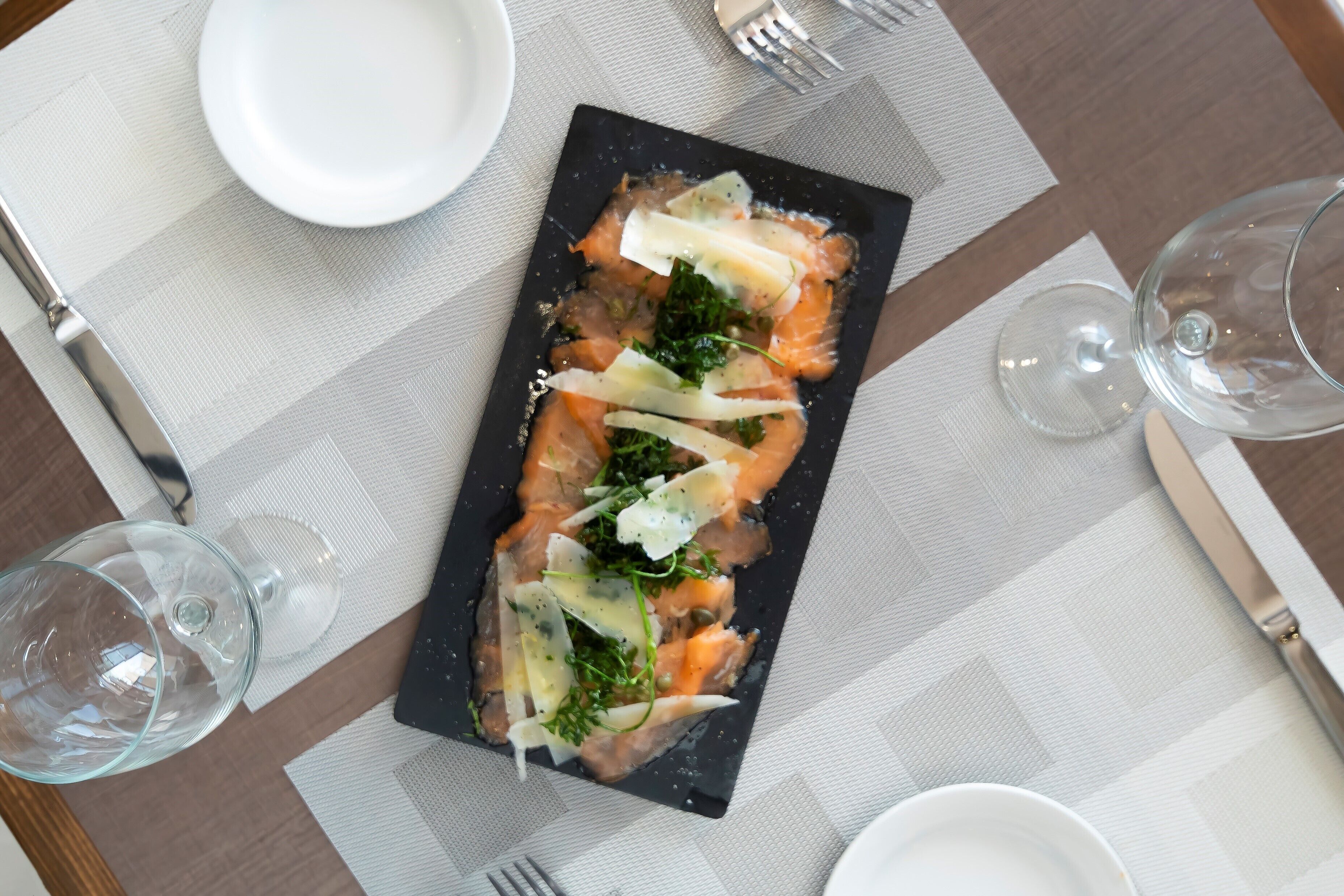Una mesa de comedor ordenada con un plato rectangular negro de comida, una copa de vino y cubiertos.
