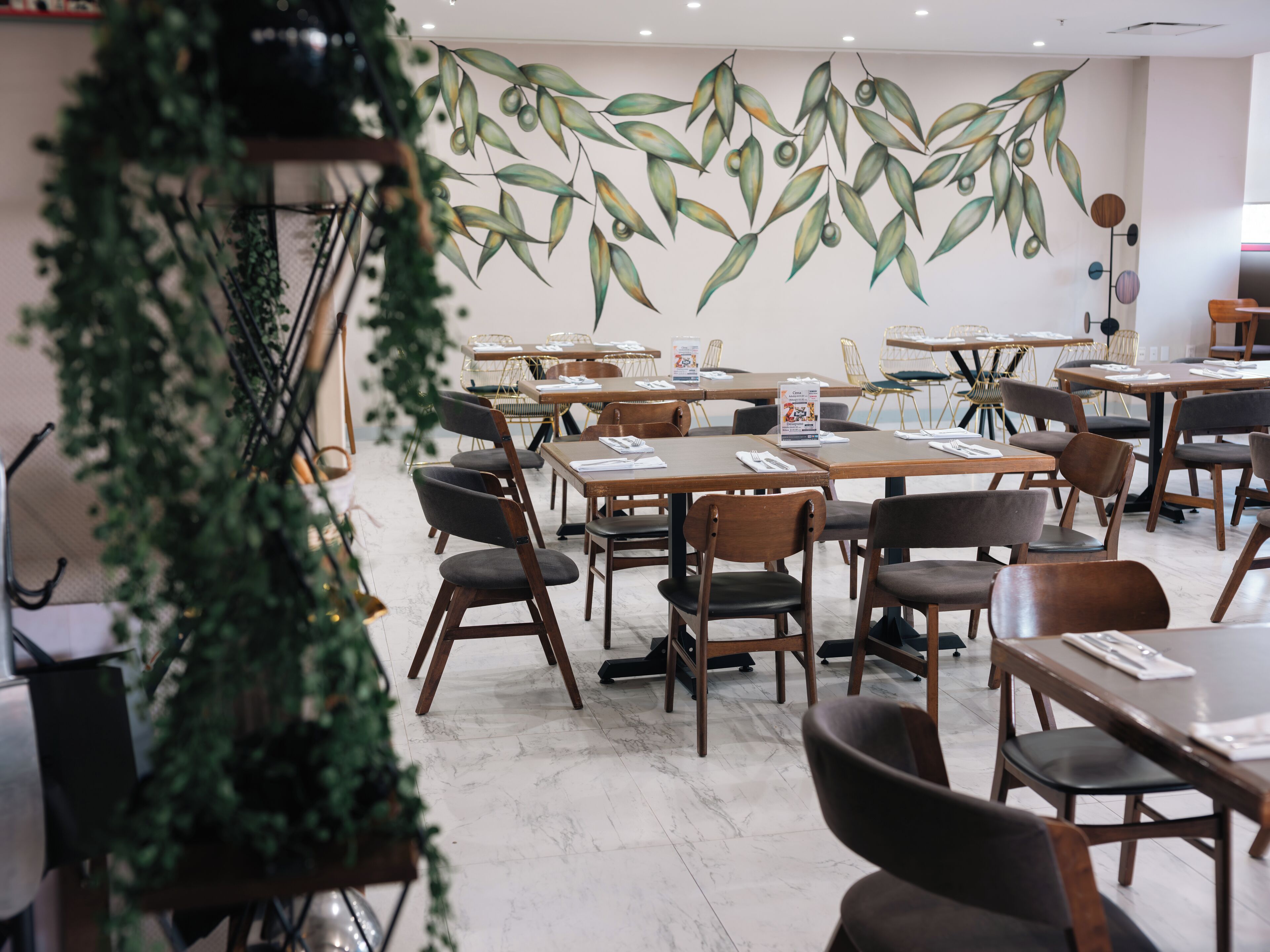 A restaurant with multiple tables and chairs, a mural of leaves on the wall, and a plant in the foreground.