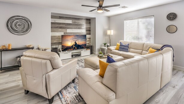 Ein modernes Wohnzimmer mit einem Ecksofa, einem Flachbildfernseher, einem Deckenventilator und einem großen Fenster mit Jalousien.