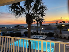 Zona de piscina con vista al atardecer sobre el océano, palmeras y un estacionamiento.