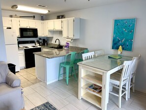 Cocina moderna con gabinetes blancos, estufa negra y encimera de granito. Hay una zona de comedor con mesa de vidrio y sillas blancas.