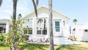 Una casa blanca con ventanas grandes, un patio con sillas y un árbol pequeño en el jardín delantero.