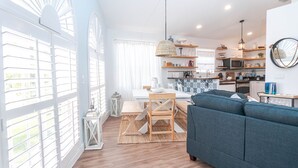 Un salón luminoso con un sofá azul, una mesa de comedor de madera y una cocina al fondo.