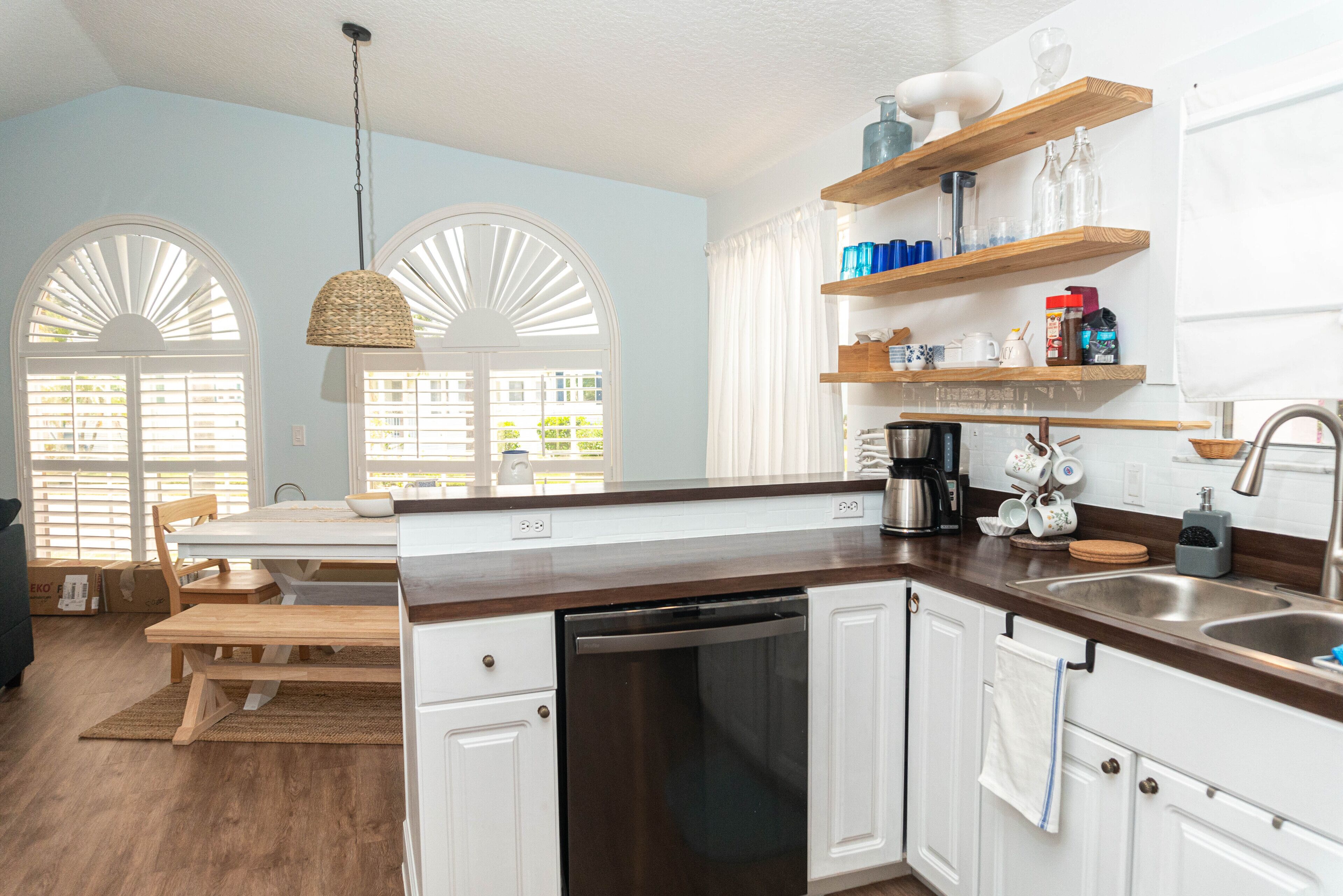 Una cocina con armarios blancos, una encimera de madera y un lavavajillas. Hay dos ventanas con contraventanas blancas, una lámpara colgante y una estantería con utensilios de cocina.