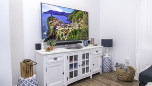Una sala de estar moderna con un televisor de pantalla plana montado en un mueble blanco, una mesita auxiliar con lámpara y una zona de estar acogedora con un sofá de color oscuro.