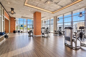 A spacious gym with various exercise machines, large windows, and a brick accent wall.
