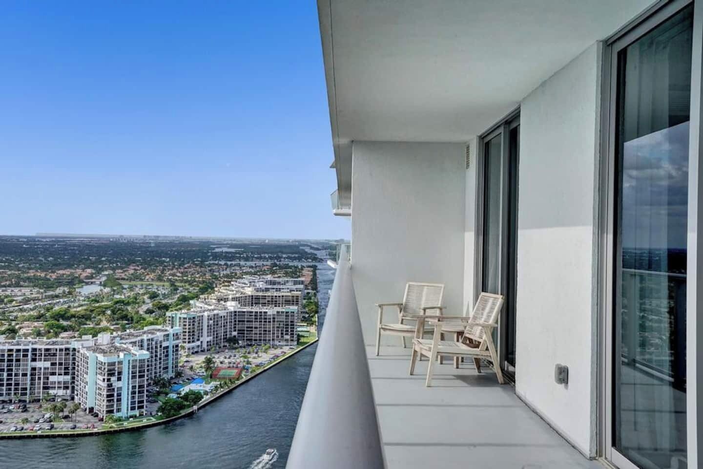 Un balcon donnant sur une rivière et des immeubles, deux chaises blanches et une porte vitrée.