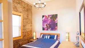 Ein Schlafzimmer mit einer Ziegelwand, einem großen Bett mit blauer Bettwäsche, einem hölzernen Kopfteil, einer Chandelieber, einem Nachttisch mit Lampe und einem Wandbild mit rosa Blumen.