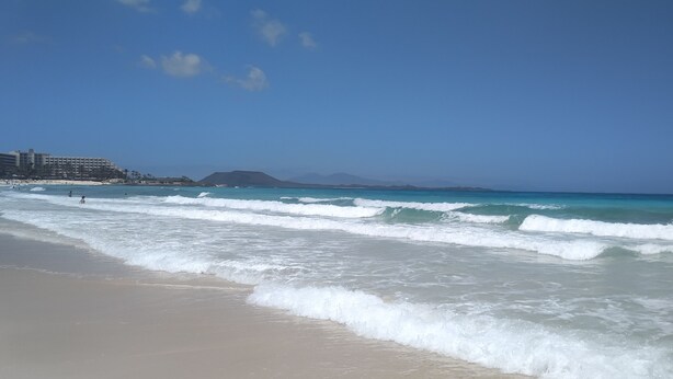 Una playa con olas, cielo despejado y montañas a lo lejos.