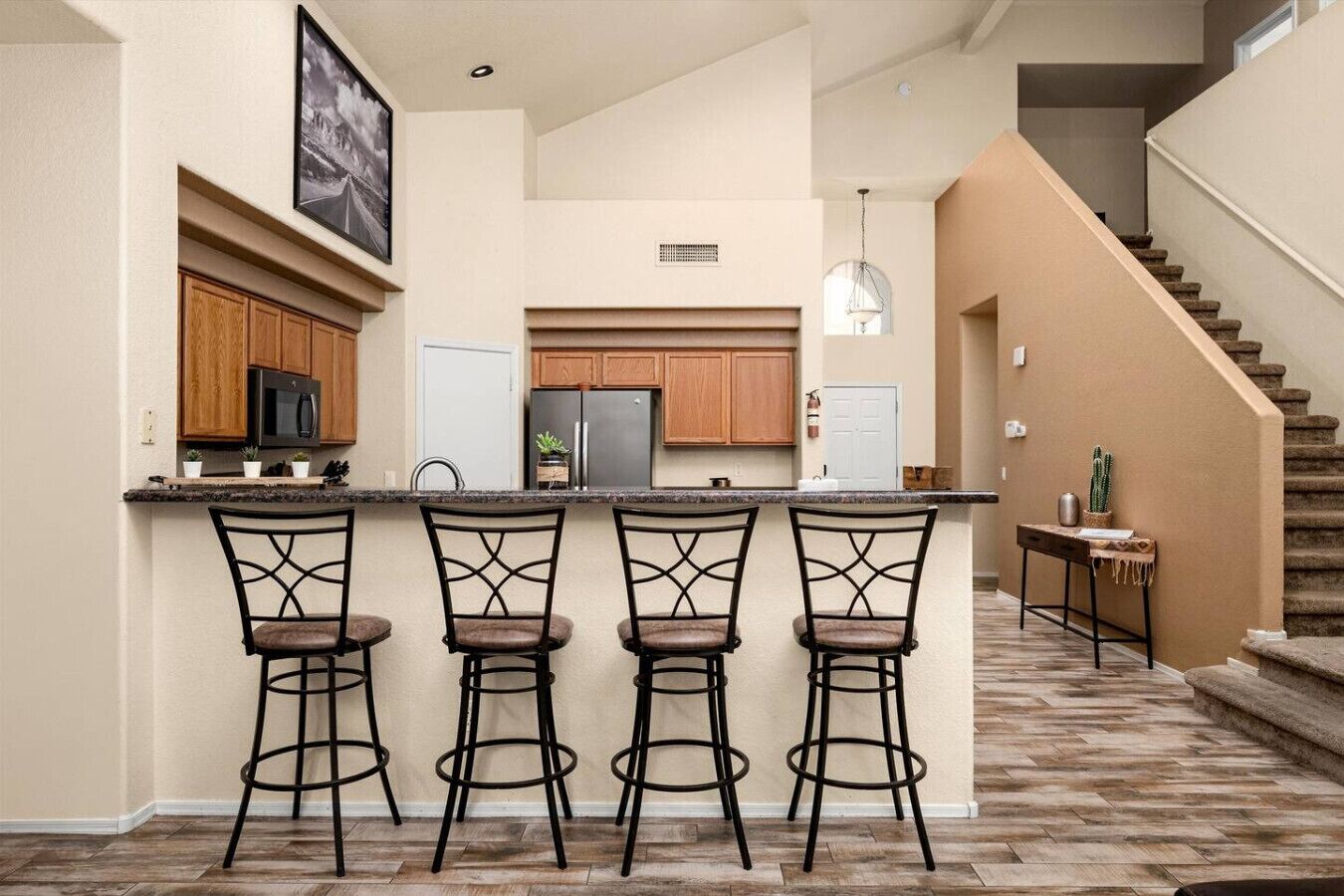 A modern kitchen with a central island, bar stools, wooden cabinets, and a staircase leading to another level.