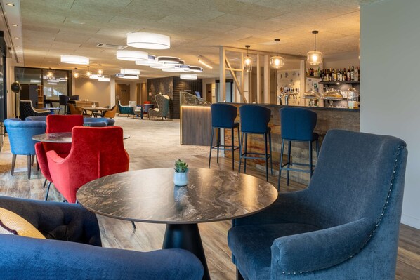 A modern lounge area with round tables, blue and red chairs, and a bar with a variety of bottles. - Best Western Plus Le Conquerant Rouen Nord (Bois-Guillaume)