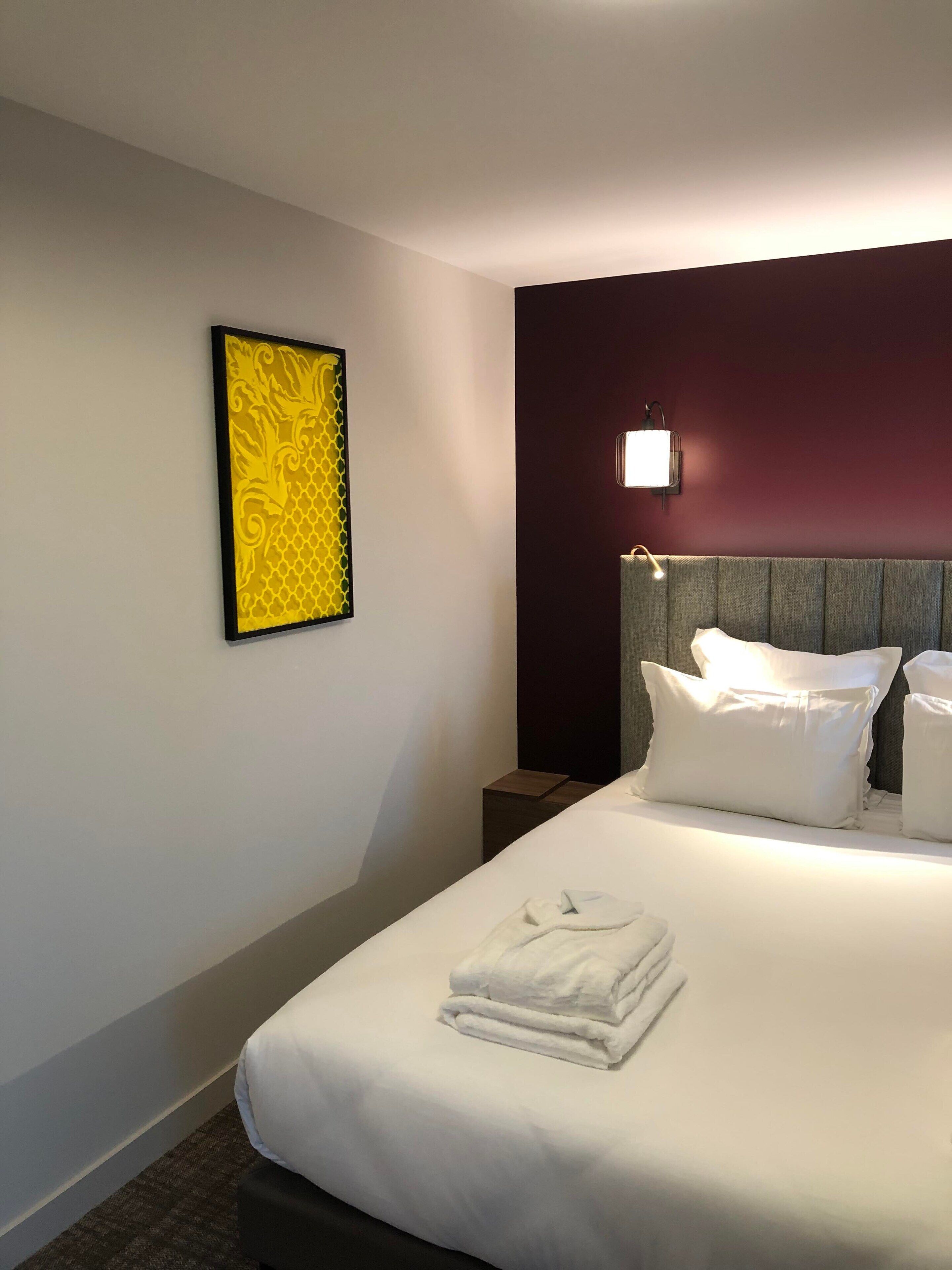 A hotel room with a neatly made bed, a nightstand, a wall-mounted lamp, and a framed artwork.