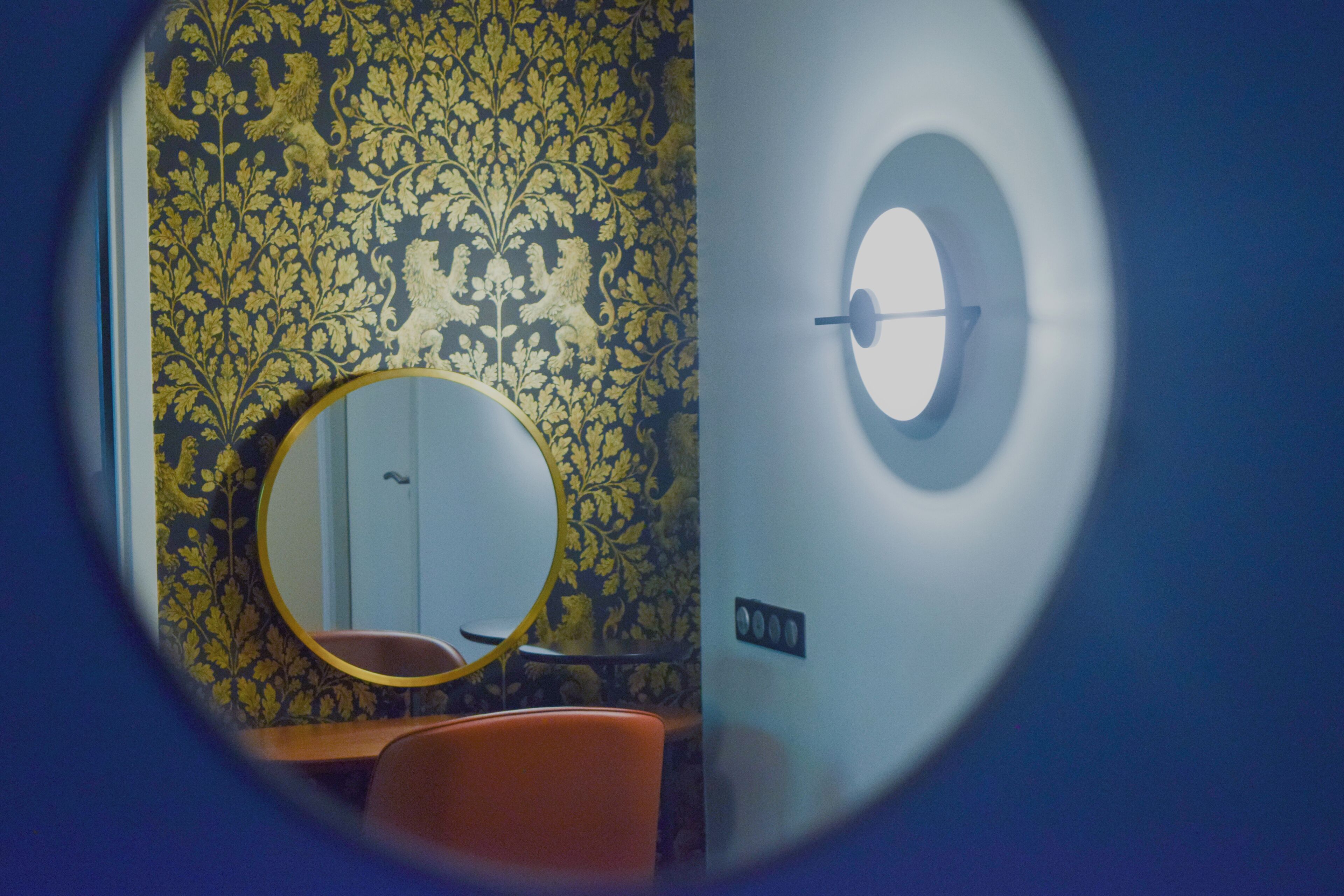 A room with a patterned wallpaper, a round mirror, a small table, and a wall-mounted light fixture.