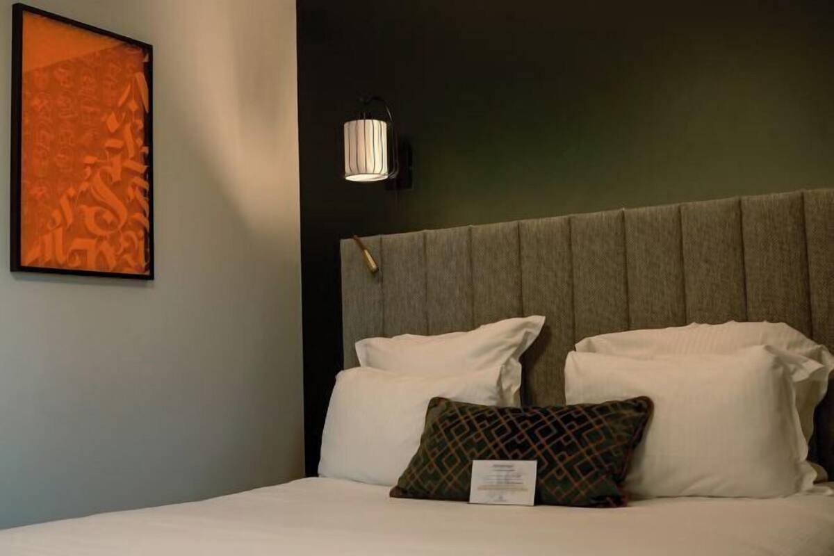 A hotel room with a bed, pillows, a wall-mounted light fixture, and a framed artwork on the wall.