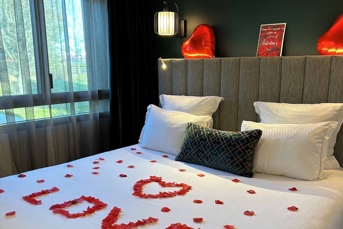 A hotel room with a bed adorned with rose petals spelling "LOVE," a nightstand with a lamp, and a view of greenery through the window.