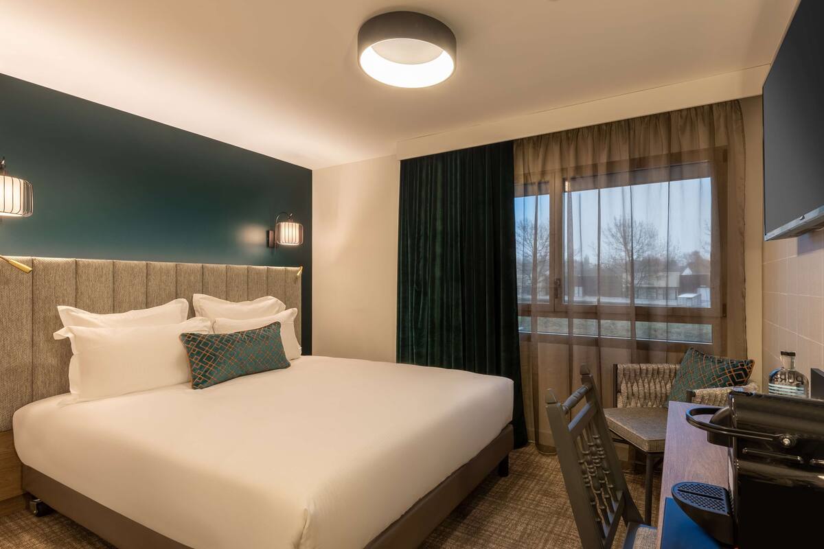 A hotel room with a large bed, a desk with a chair, a window with curtains, and a wall-mounted lamp.