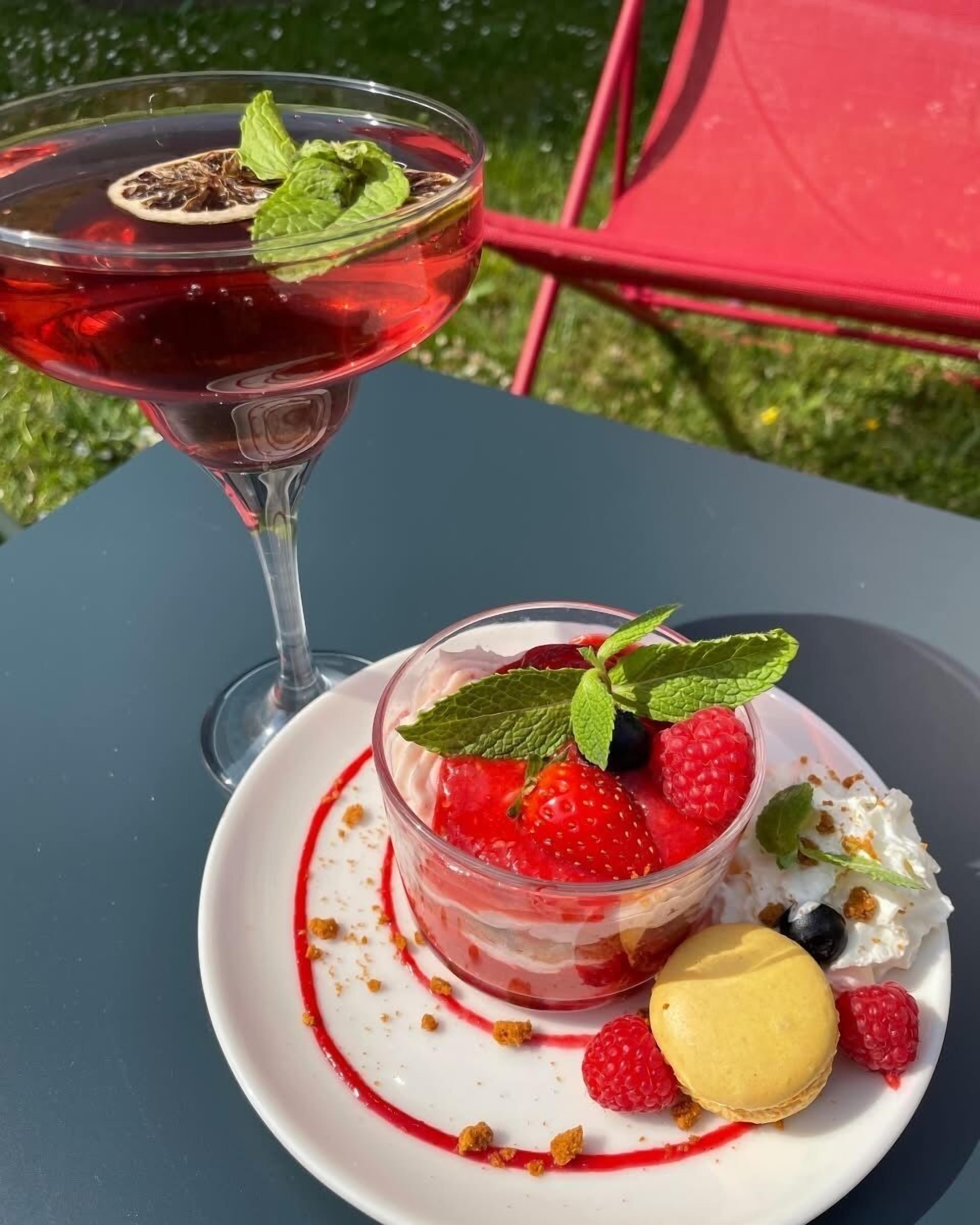 Un dessert aux fraises et aux framboises, avec un élément jaune sur une assiette blanche, et un verre d’une boisson rouge décorée de feuilles de menthe, le tout sur une table à l’extérieur.