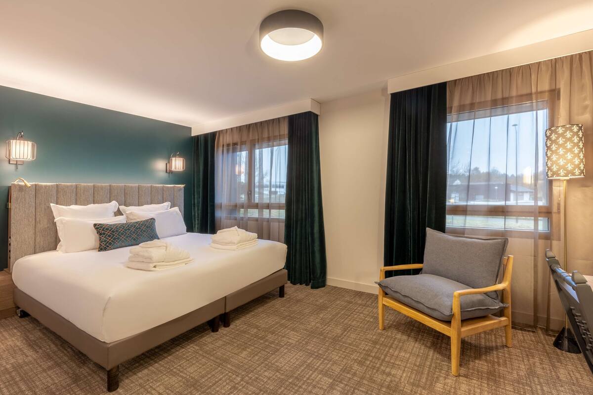 A hotel room with a large bed, a chair, a desk, and a window with curtains.