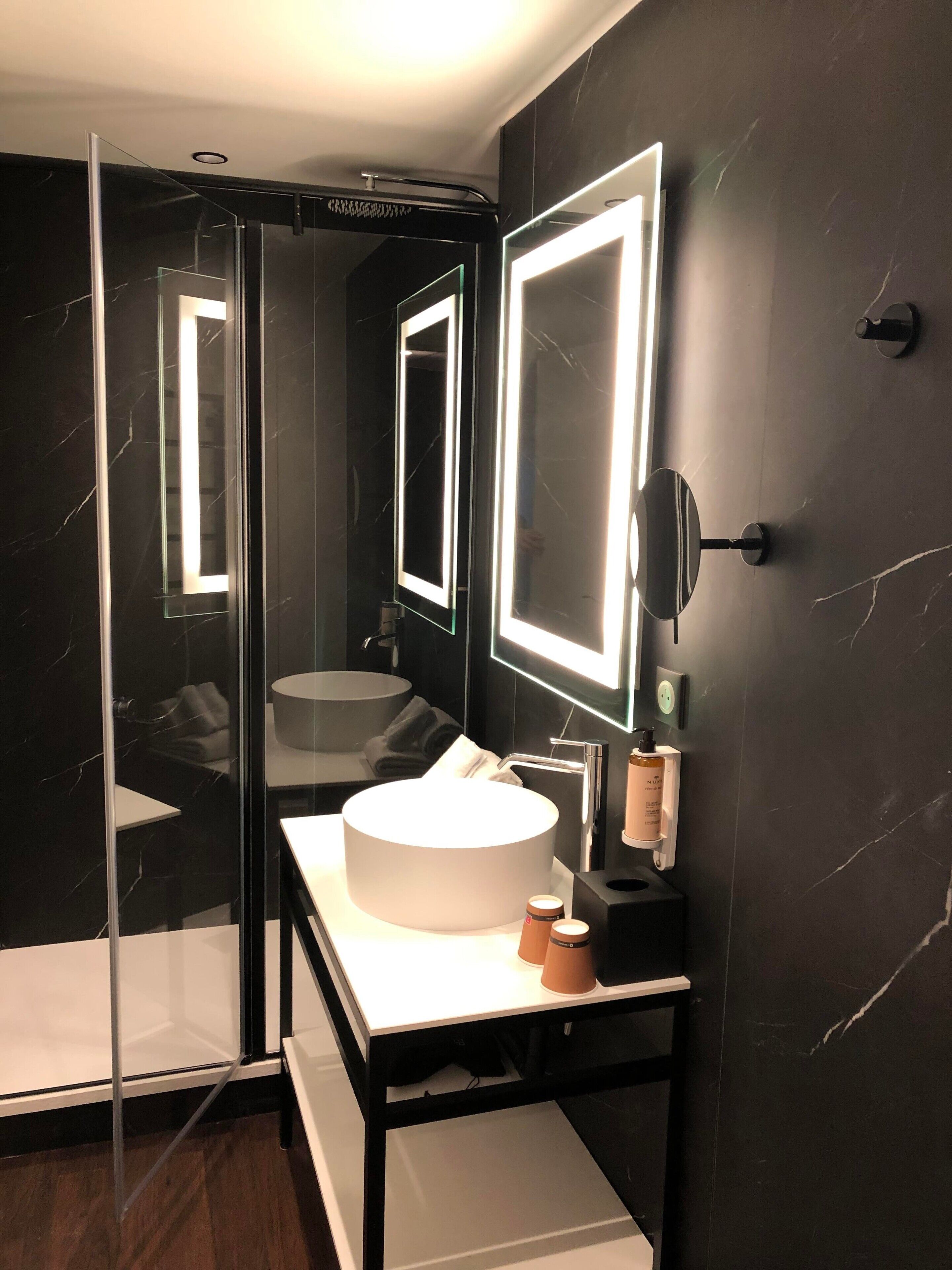 A modern bathroom with a round sink, a glass shower enclosure, and a large mirror with integrated lighting.
