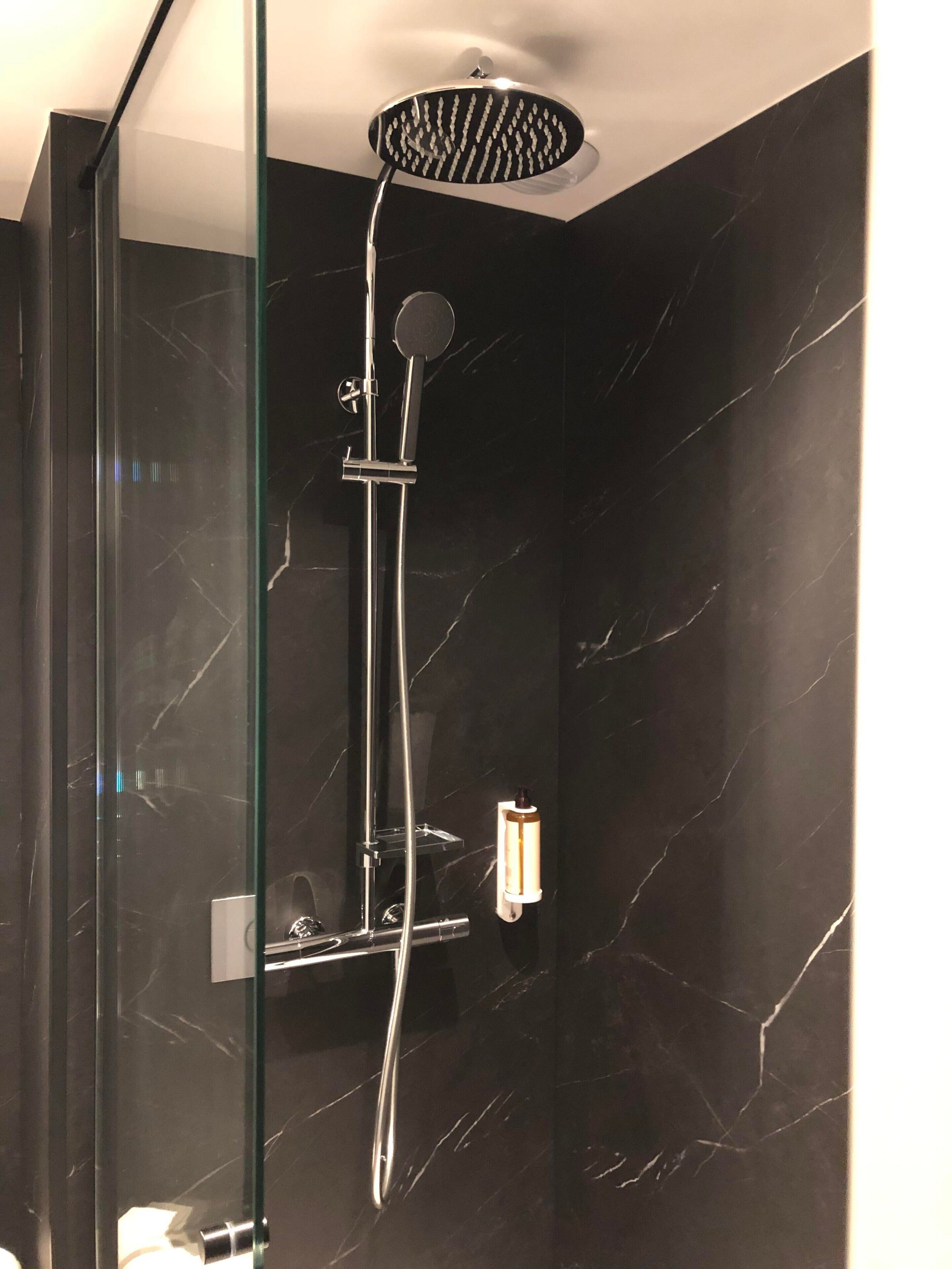 Une douche moderne dotée d’un pommeau de douche à pluie et d’un pommeau de douche à main, le tout installé sur un mur en marbre foncé.