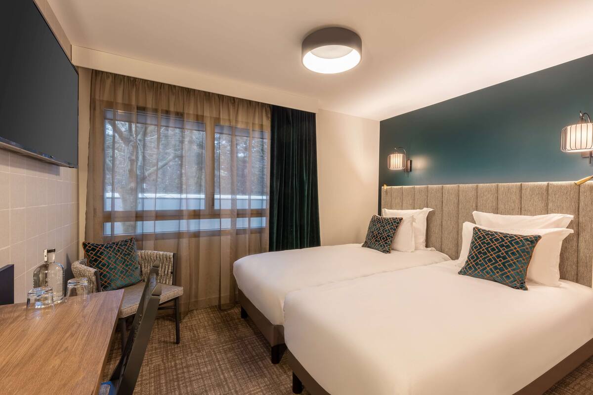 A hotel room with two beds, a desk with a chair, a large window with curtains, and a wall-mounted light fixture.