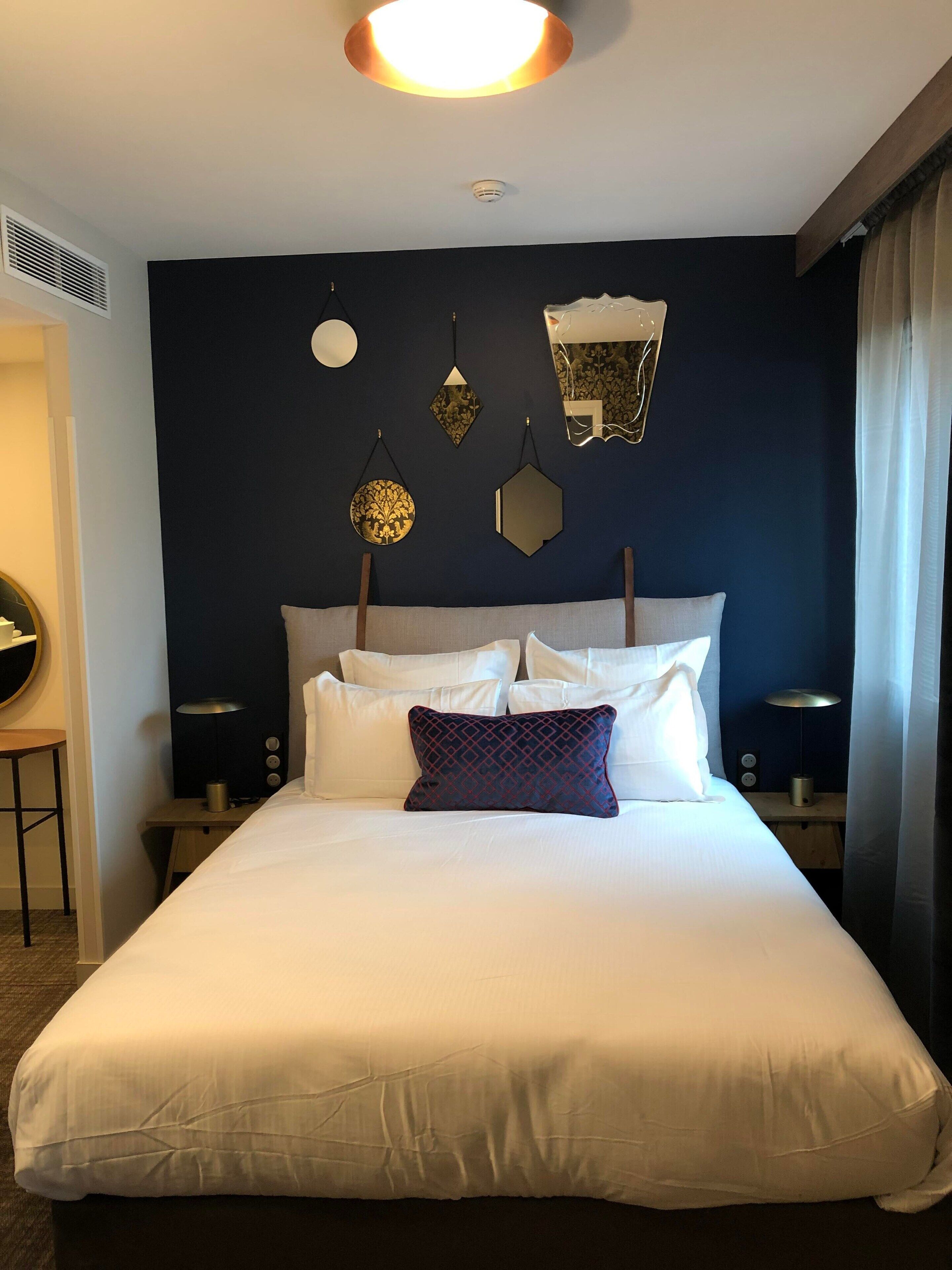 Ein Hotelzimmer mit einem großen Bett, weißer Bettwäsche und einer dunkelblauen Akzentwand. An den Wänden sind Wandleuchten angebracht, eine Deckenleuchte strahlt herab und ein Nachttisch mit einer Lampe steht bereit.