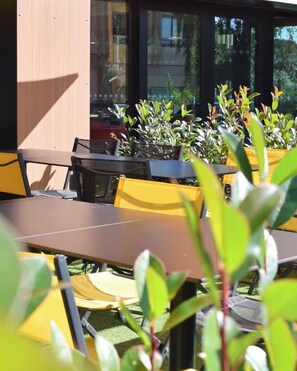 An outdoor seating area with tables and chairs, surrounded by potted plants, and a building with large windows in the background. - Best Western Plus Le Conquerant Rouen Nord (Bois-Guillaume)