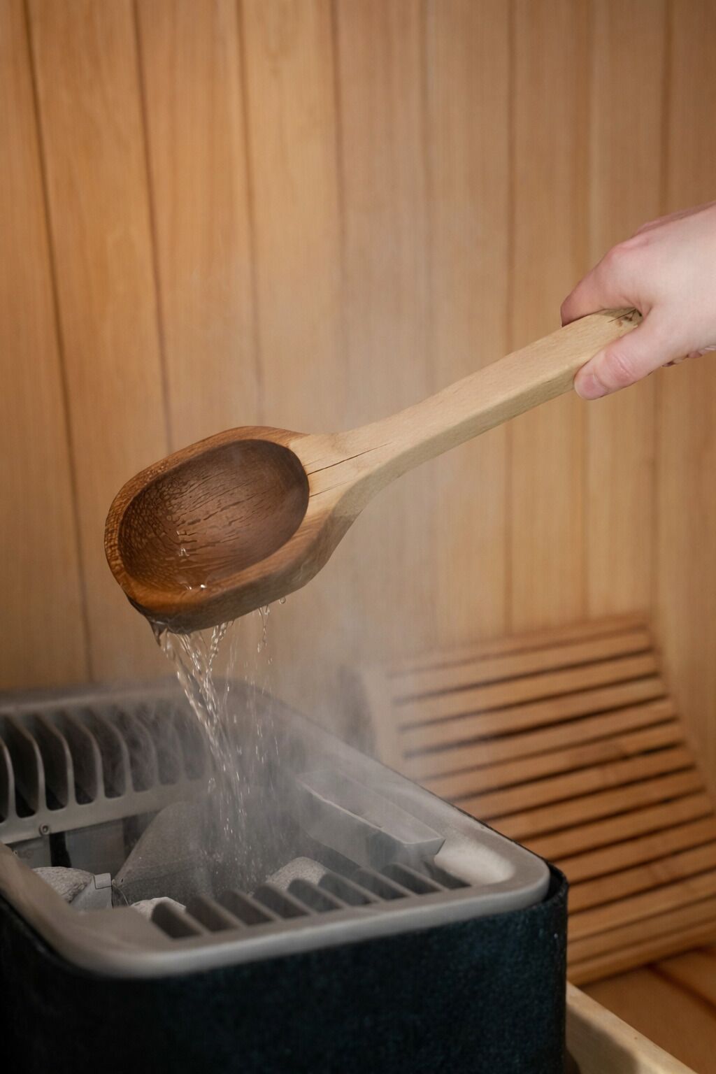 Una mano sostiene una cuchara de madera vertiendo agua en una sauna.