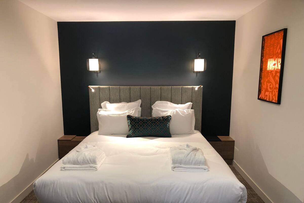 A neatly made bed with white linens and two pillows, a headboard with integrated lighting, and a framed artwork on the wall.