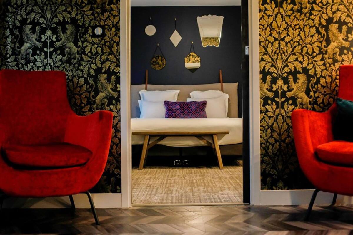 A room with a bed, two red chairs, a patterned wallpaper, and a mirror on the wall.
