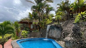 Una piscina con cascata, circondata da vegetazione tropicale e un muro in pietra.