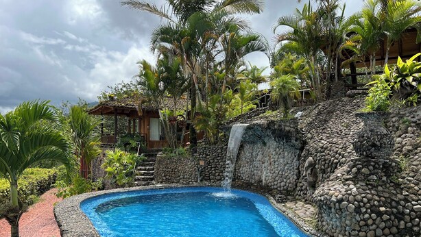 A swimming pool with a stone wall and a waterfall, surrounded by tropical vegetation. - Wide Luxury Home with Pool & Jacuzzi access & Amazing Volcano and Lake views (El Castillo)