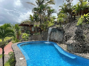 Une piscine bordée d’un mur en pierre et agrémentée d’une cascade, le tout entouré d’une végétation tropicale.