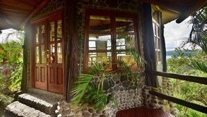 A stone and wood building with a wooden deck, a stone staircase, and a view of the surrounding greenery and landscape. - Wide Luxury Home with Pool & Jacuzzi access & Amazing Volcano and Lake views (El Castillo)