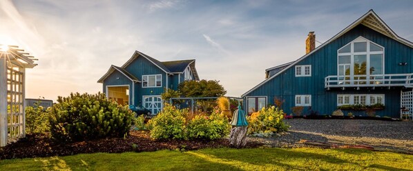A large, two-story house with a blue exterior, a visible garage, and a well-maintained garden with various plants and flowers. - Waterfront Studio Private Guest Suite at Dungeness Barn House (Sequim)
