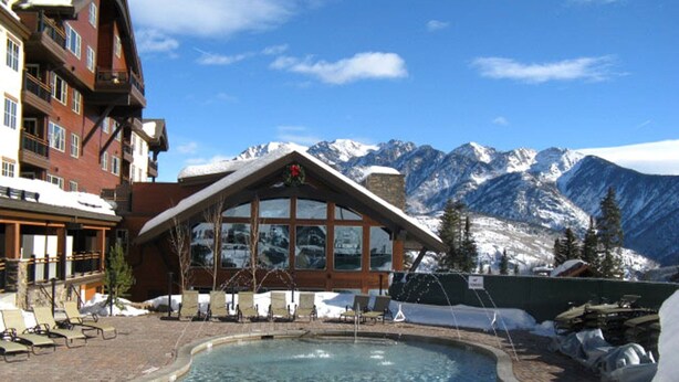 Ett hotell med en pool omgiven av snö, utsikt över ett berg och en byggnad med stora fönster.