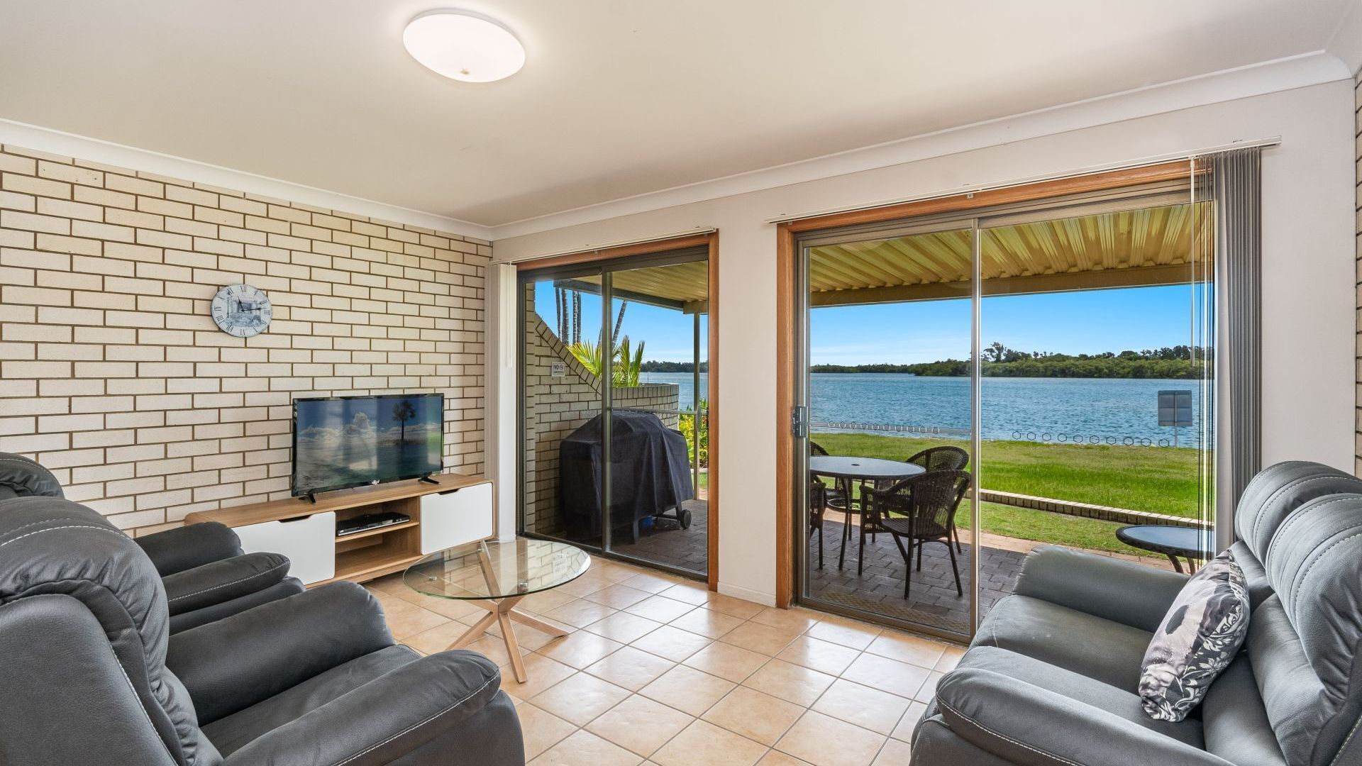 A modern living room with a TV, sofa, and a glass table. It features a brick wall, a sliding glass door leading to a balcony with a view of a lake.