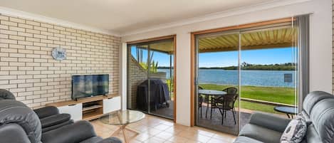 A modern living room with a TV, sofa, and a glass table. It features a brick wall, a sliding glass door leading to a balcony with a view of a lake.