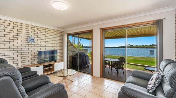 A modern living room with a TV, sofa, and a glass table. It features a brick wall, a sliding glass door leading to a balcony with a view of a lake.