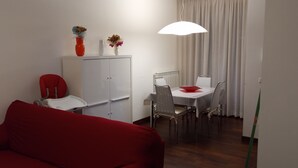 Een knus woongedeelte met een rode bank, een eettafel met stoelen, een witte kast en een hanglamp.